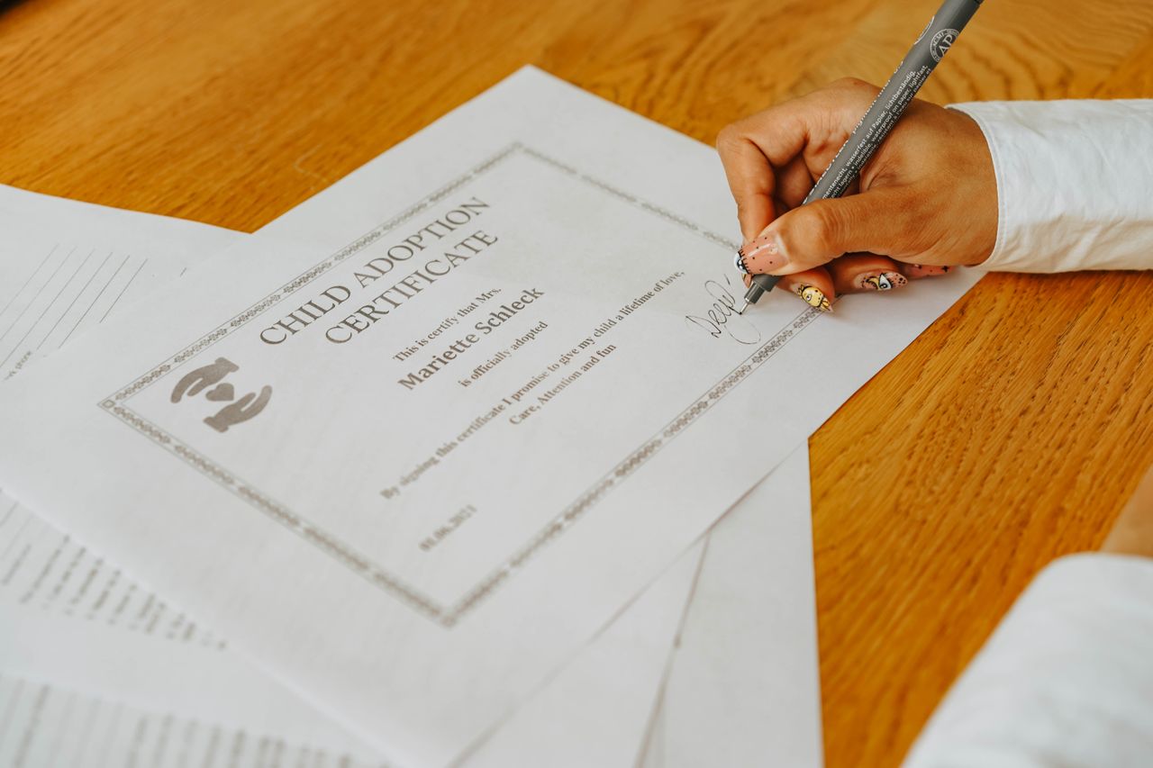 Services A close-up of a hand signing a child adoption certificate with a pen on a wooden table.